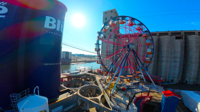 Colorful ferris wheel near water with buildings and a bright sun in the background.