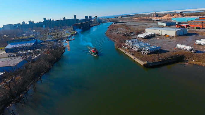 Aerial view of a river with a boat, surrounded by industrial buildings and trees.